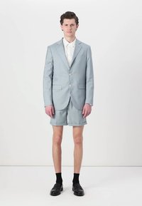 Completo azzurro chiaro con giacca monopetto e pantaloni corti abbinati. Presenta una vestibilità su misura, una texture morbida e un paio di mocassini neri con calze.