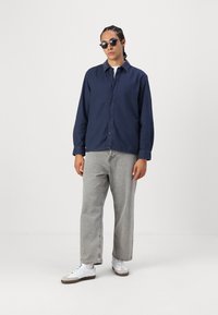 Navy button-up shirt with a relaxed fit and long sleeves, paired with light gray straight-leg jeans and white sneakers. Black sunglasses worn.