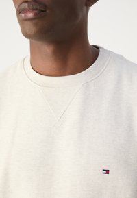 Nahaufnahme einer Person, die einen hellbeigen Sweatshirt mit einem kleinen Tommy Hilfiger-Flaggenlogo nahe der Brust trägt.