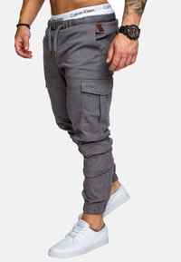 Graue Cargo-Jogginghosen mit elastischen Bündchen und einem Kordelzugbund, ausgestattet mit Seitentaschen und einem Lederakzent. Getragen mit weißen Sneakers.