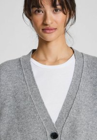 Cardigan grigio a maglia con scollo a V, caratterizzato da un motivo testurizzato e un unico bottone nero. Indossato sopra una maglietta bianca.