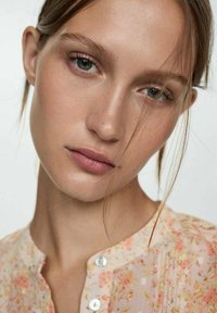 Licht bloemenblouse met een knoopdetail, met zachte roze tinten en subtiele texturen. Het haar valt zachtjes langs het gezicht.