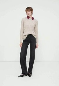 Frau mit kurzen Haaren, die einen beigen Pullover mit burgunderfarbenem Kragen, schwarze hoch taillierte Jeans und schwarze spitze Schuhe trägt, steht vor einer weißen Wand.