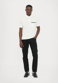 Hvit kortermet polo skjorte med lomme og logo i svart, kombinert med svarte slim-fit bukser og svarte loafers. Ensfarget bakgrunn.