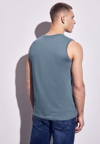 Tyrkysový top bez rukávů vyrobený z lehké látky, s kulatým výstřihem, mírně uvolněným střihem a měkkou texturou, doplněný modrými džíny.