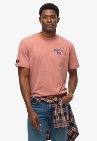 Camiseta de algodón rosa con mangas cortas y un estampado gráfico azul. El modelo lleva unos vaqueros de mezclilla y una camisa a cuadros atada a la cintura.