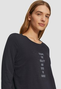 Schwarz langärmliges T-Shirt mit rundem Ausschnitt, verziert mit weißen Texten und kleinen silbernen Nieten, die in einem rechteckigen Muster auf der Vorderseite angeordnet sind.