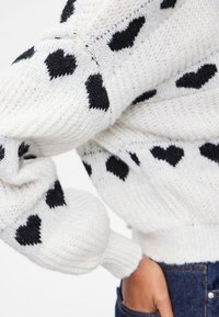 Vit stickad tröja med svarta hjärtan, ribbade ärmmuddar och en texturerad yta. Designen ger en mysig och avslappnad look.