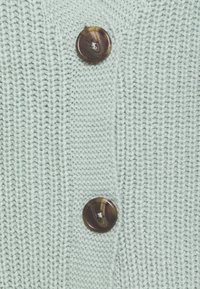 Jasnozielony sweter z dzianiny z teksturowanym wzorem, z dwoma okrągłymi brązowymi guzikami z przodu do zapięcia.