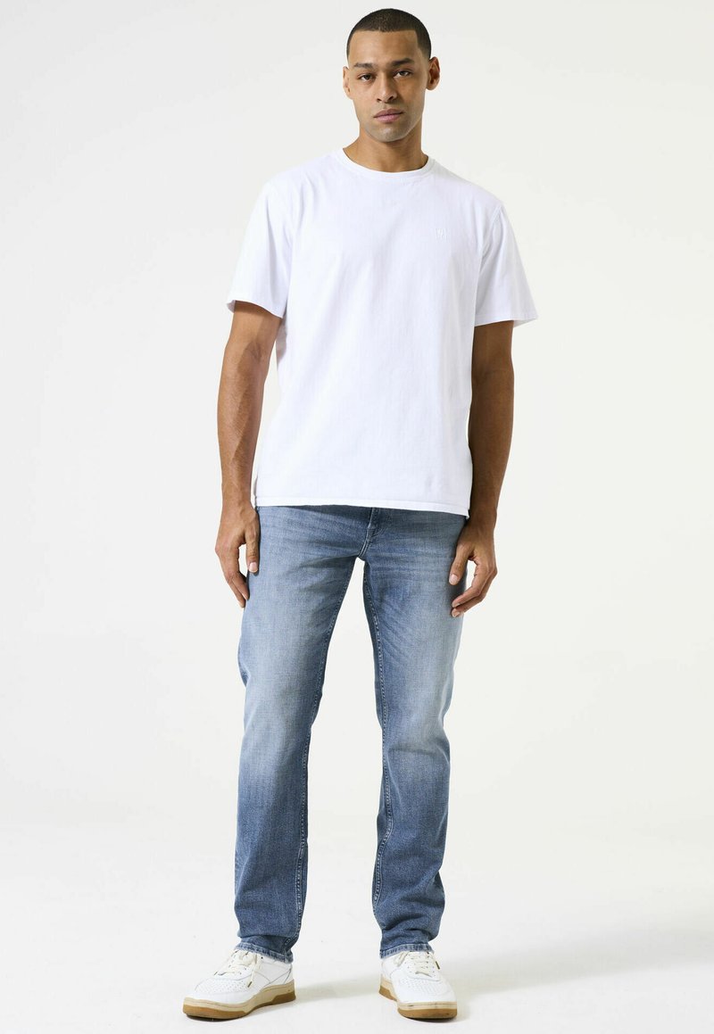 Camiseta de algodón blanca con cuello redondo combinada con unos vaqueros de mezclilla azul claro. Zapatillas blancas con acentos texturizados y suelas marrón claro.