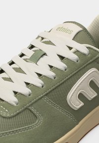 Πράσινο και μπεζ αθλητικό παπούτσι Etnies με λευκά κορδόνια, ορατό λογότυπο στο πλάι, υφές από δίχτυ και σουέντ, και ετικέτα με την επωνυμία στη γλώσσα του παπουτσιού.
