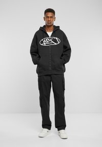 Sort zip-up hættetrøje med hvidt logo på brystet, parret med sorte cargo bukser med sidelommer og hvide sneakers. Enkel, afslappet pasform.