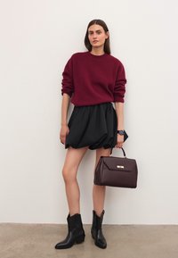 Mujer vestida con un suéter burdeos y una falda negra abullonada, sosteniendo un bolso marrón oscuro, de pie contra una pared clara y lisa.