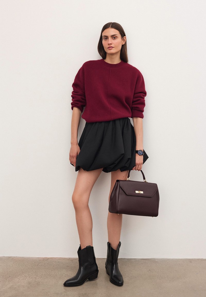 Mujer vestida con un suéter burdeos y una falda negra abullonada, sosteniendo un bolso marrón oscuro, de pie contra una pared clara y lisa.