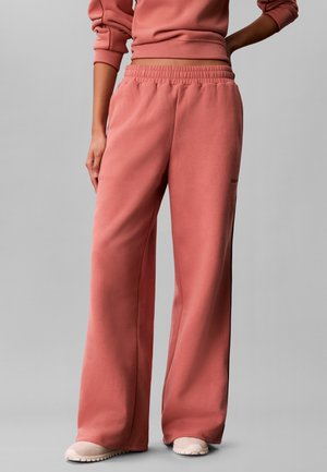 Person iført løstsiddende, højtaljede, støvet rosa joggingbukser og matchende cropped sweatshirt, stående på lysegrå baggrund.