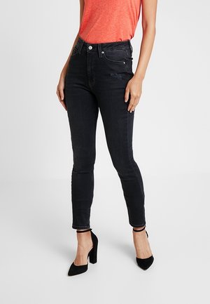 Femme portant un jean skinny noir taille haute, un haut orange à manches courtes et des chaussures à talons noirs avec bride à la cheville, debout sur fond blanc.