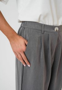 Sive krojene hlače s sproščenim krojem, ki vključujejo stranske žepe in gumbasto zapiranje. Tkanina ima gladko teksturo in subtilen lesk.