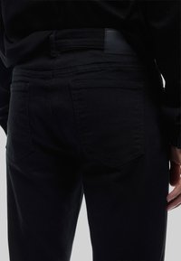 Jeans de denim negro con un diseño ajustado, bolsillos traseros con solapas en ángulo y una textura suave. La cintura tiene un pequeño parche de logo negro.