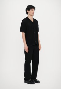 Juodas trumpas rankovės mygtukinis marškinėlis su tekstūruotu megzto dizainu, derinamas su juodomis tiesiomis kelnėmis ir blizgiais juodais batais.