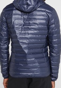 Marinblå pufferjacka med en glänsande, quiltad textur. Den har en huva, horisontell söm och elastiska ärmslut för en åtsittande look.