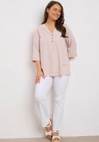 Lächelnde Frau, die an einer weißen Wand lehnt und eine blassrosa geknöpfte Bluse, weiße Hose und weiße Keilsandalen trägt.