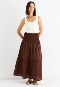 Brauner, stufenförmiger Maxirock mit gesmocktem Bund, kombiniert mit einem weißen Tanktop. Das Model trägt braune Sandalen. Glatte Stoffstruktur, lässiges Design.