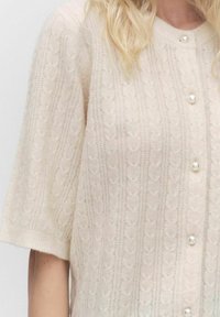Světle béžový pletený kardigan s texturovaným vzorem, krátkými rukávy a kulatým výstřihem. Obsahuje dekorativní perlové knoflíky vpředu.
