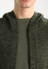 Felpa con cappuccio verde oliva in tessuto testurizzato, progettata con apertura frontale e uno strato interno morbido. La texture presenta un motivo sottile.