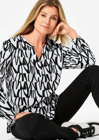 Zwart-wit geblokte blouse met een V-hals en een placket met twee knopen; lange mouwen met elastische manchetten, gedragen met zwarte broek en meerkleurige schoenen.