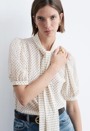 Mujer con blusa crema de estampado pequeño y mangas abullonadas, combinada con jeans azules y pendientes de aro dorados y anillo.