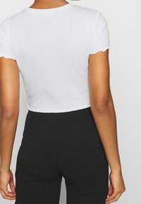 Osoba ubrana w dopasowany biały krótki top z krótkim rękawem i czarne spodnie z wysokim stanem, pokazana od tyłu na jednolitym tle.