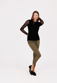 Zwart langwerpig shirt met doorschijnende mesh mouwen, gecombineerd met olijfgroene skinny jeans en zwarte gepatineerde instapschoenen.