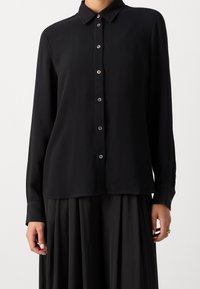 T-shirt nero a maniche lunghe con chiusura a bottoni sul davanti, colletto classico, trama a coste e tessuto liscio. I dettagli includono bottoni scuri e polsini.