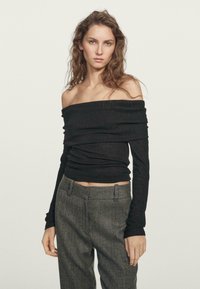 Schwarzes, schulterfreies Langarmoberteil mit strukturiertem Finish, kombiniert mit grauen, hochgeschnittenen Hosen, die über ein dezentes Karomuster verfügen.