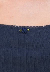 Tumši zils adīts tops ar ribotiem rakstiem un smalku mežģīņu apdari. Vēl ir neliela auduma roze tumši zilā un zaļā krāsā ap kakla līniju.