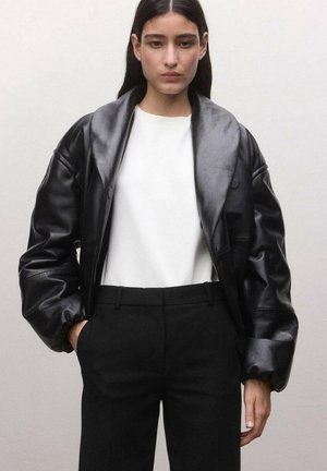 Mujer con una chaqueta de cuero negra extragrande, camiseta blanca y pantalones negros, con las manos en los bolsillos, sobre un fondo liso.