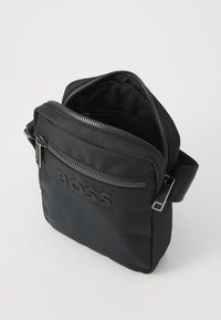 Borsa a tracolla nera in nylon con tracolla regolabile, tasche con zip e logo impresso sul davanti. L'interno sembra spazioso con una texture liscia.