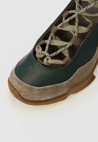Green leather shoe with a textured brown suede toe cap, featuring braided laces in beige and brown, and a ridged rubber sole.