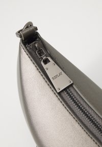 Pochette grigia metallica con una superficie liscia, caratterizzata da una cerniera nera e un'etichetta in metallo argentato contrassegnata "REPLAY". Design compatto e elegante.