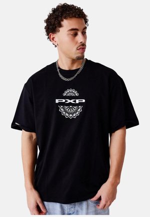 Joven con pelo rizado que lleva una camiseta negra con texto blanco "PXP" y diseño geométrico, cadena de plata y vaqueros azul claro.