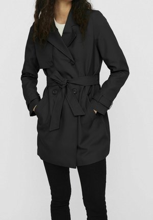 Mujer con un abrigo trench negro de doble botonadura con cinturón anudado en la cintura y manos en los bolsillos, de pie frente a un fondo claro y liso.