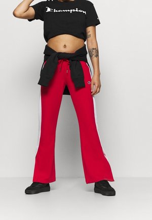 Pantalones deportivos rojos acampanados con rayas blancas laterales y cintura con cordón, combinados con una camiseta negra corta, usados con zapatillas negras.