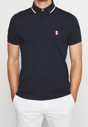 Navy blue polo shirt with a small red and white logo, featuring a contrasting white collar and short sleeves, made from smooth fabric.