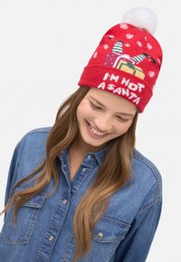 Gorro de punto rojo con un pompón blanco, con diseños de regalos coloridos y el texto "NO SOY SANTA." El fondo está decorado con patrones de copos de nieve.