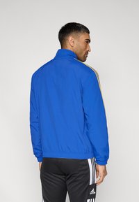 Blue track jacket with a high collar, elastic waistband, and gold stripes along the shoulders, paired with black athletic pants.