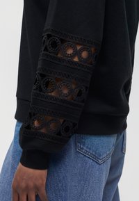 Haut noir à manches longues avec des découpes en dentelle circulaires sur la manche, porté avec un jean en denim bleu clair sur la partie inférieure du corps.