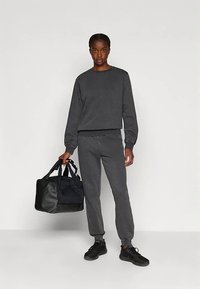 Sieviete stāv tumši pelēkā sporta krekliņā un sporta biksēs, kreisajā rokā tur melnu sporta somu, pret vienlīdzīgu gaišu fona.