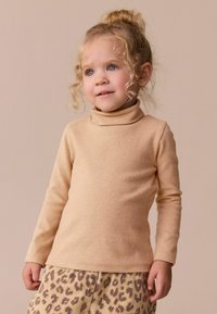 Młoda dziewczyna z kręconymi, blond włosami, ubrana w beżowy sweter z golfem i spódniczkę w panterkę, patrząca lekko na bok z delikatnym uśmiechem.