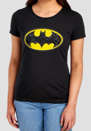 Frau trägt ein schwarzes T-Shirt mit einem großen gelb-schwarzen Batman-Logo vorne und hellblaue Jeans.