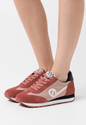 Pieds portant des baskets rose et rouille avec un logo blanc et une étiquette à carreaux sur la languette, debout sur un fond blanc.